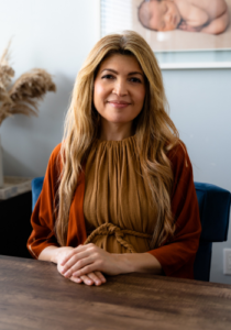 The image is of Ivelisse DeJongh, founder and lead consultant at Ruhe Fertility and Prenatal. She is a woman with long hair sitting at a desk with her hands folded in front of her.